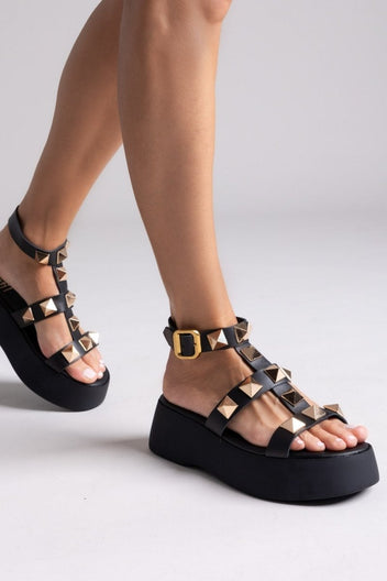 Model wearing black leather flatform sandals with gold pyramid studs and gold ankle buckle straps, angled view