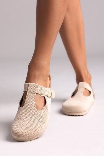 Model legs in cream suede T-strap clogs with cut-out vamp and buckle, angled view on a light backdrop