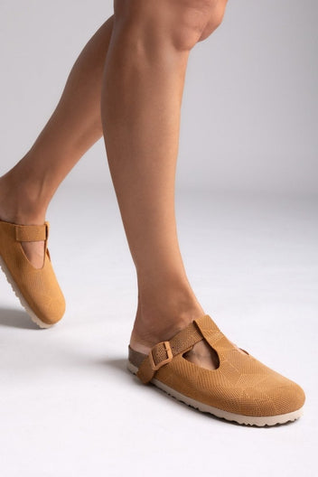Model leg in tan suede T-strap clog with cut-out vamp and buckle, angled view on a light grey backdrop