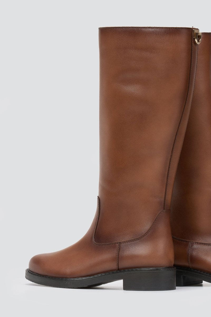 Cognac brown leather knee-high riding boot with flat sole and side zip, angled view on a light grey background