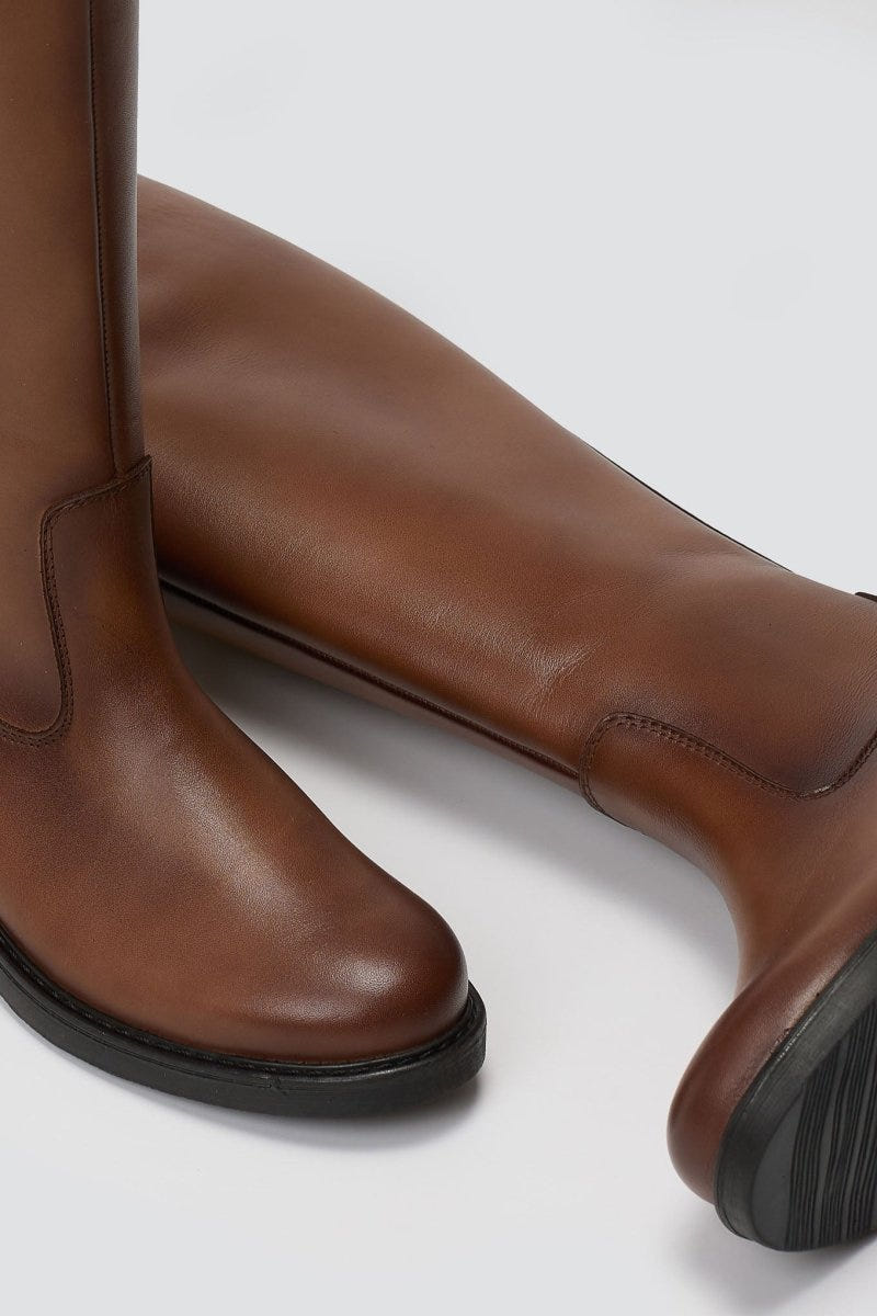 Close-up of cognac brown leather riding boot shaft and rounded toe with dark sole, detail view on light grey