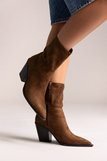 Model leg raised in dark brown suede pointed ankle boot with block heel, angled view on a light backdrop