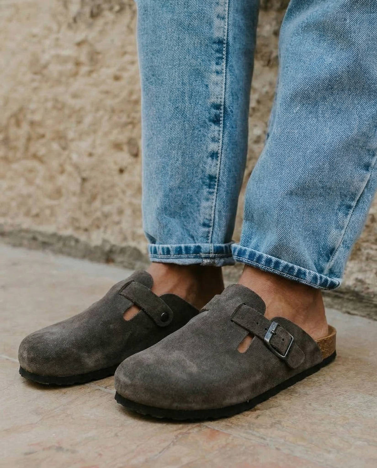 Grey suede closed-toe clogs with side buckle straps and black soles, worn with rolled-up light blue jeans on pale stone paving