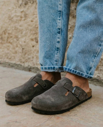 Grey suede closed-toe clogs with side buckle straps and black soles, worn with rolled-up light blue jeans on pale stone paving