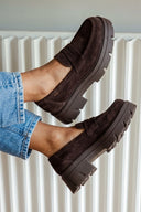 A pair of dark brown suede penny loafers with chunky lug soles, shown on a model's feet wearing blue jeans in front of a white radiator.