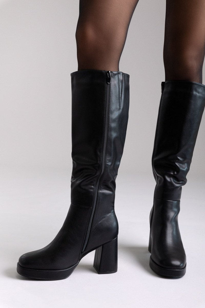 Model legs in black leather knee-high platform boots with block heels and inner side zips, front view