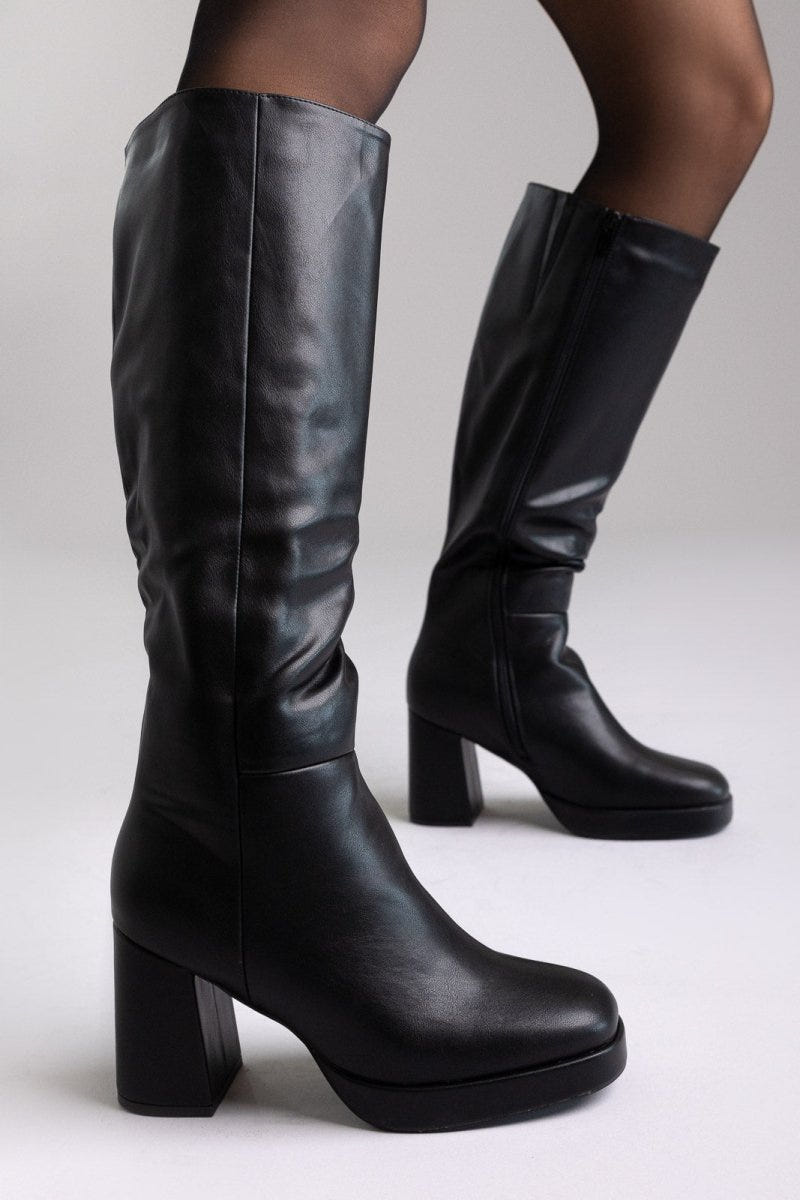Model legs in black leather knee-high platform boots with block heels and sheer tights, three-quarter view