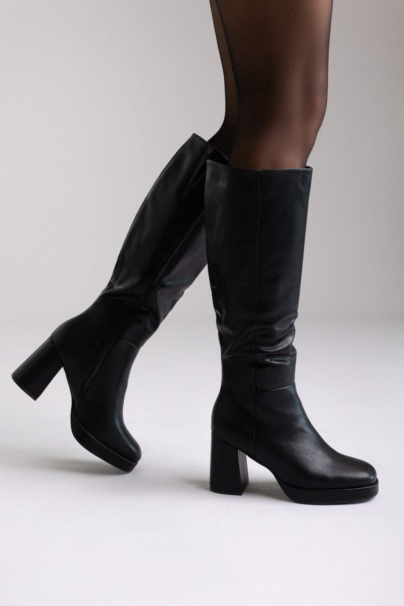 Model walking in black leather knee-high platform boots with block heels and black tights, side view