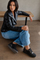 A woman sits on a wooden floor in front of a wooden bench, wearing a black leather blazer, light blue jeans, and black lace-up sneakers with silver hardware.