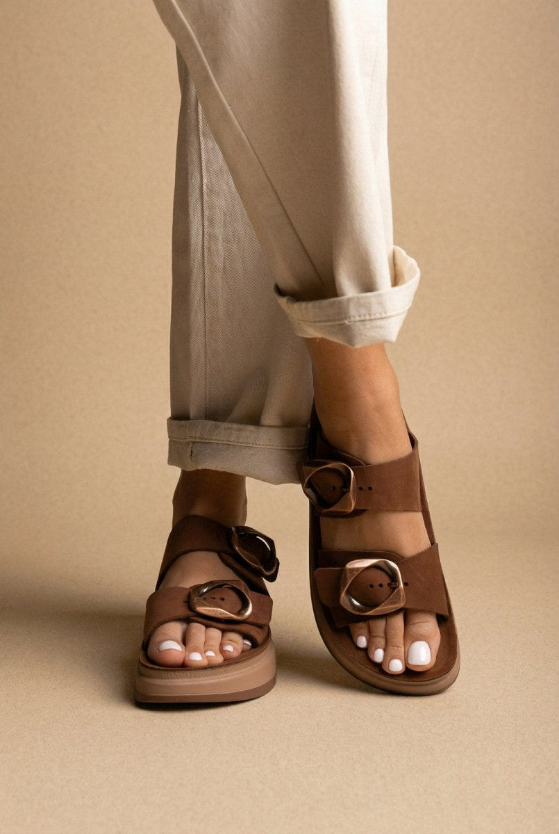 Model wearing brown suede flatform sandals with two straps and round buckles, cream rolled-cuff trousers, three-quarter view of feet