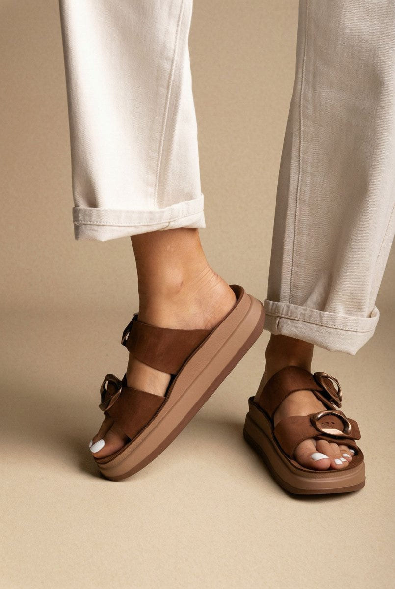 Model wearing brown suede flatform sandals with thick sole and round buckle straps, cream rolled trousers, seen from the front