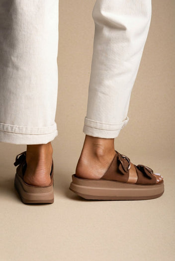 Model wearing brown suede flatform sandals with buckle straps, cream trousers cuffed, close view of feet from the side