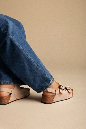 Model wearing taupe suede flatform sandals with a buckle strap, close-up of feet below cuffed jeans on a beige surface
