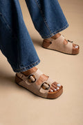 Model wearing taupe suede flatform sandals with two straps and gold ring buckles, blue jeans, three-quarter view of feet