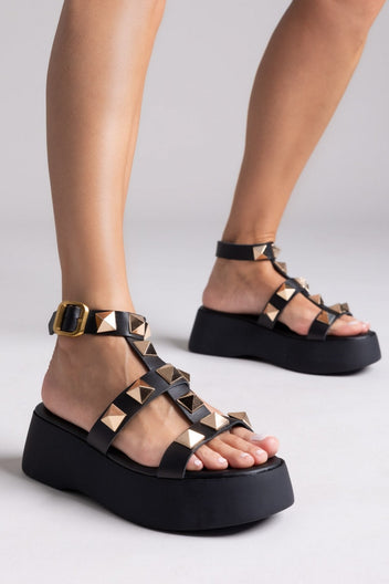 Model wearing black leather flatform sandals with gold pyramid studs and buckled ankle straps, three-quarter view