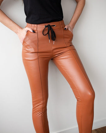Close-up of rust-orange faux leather leggings with black drawstring waist, front pockets and vertical front seams, worn on model, front view