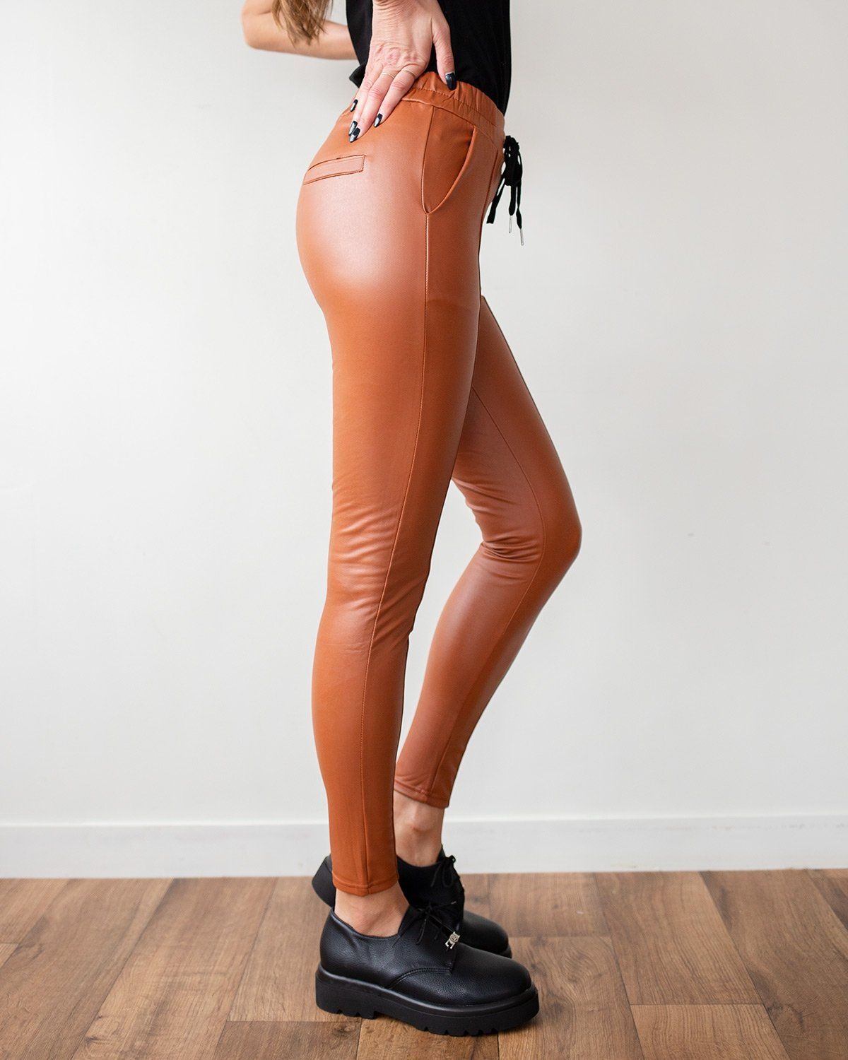 Rust-orange faux leather leggings with drawstring waist and side pocket, slim fit to the ankle, worn on model, side view