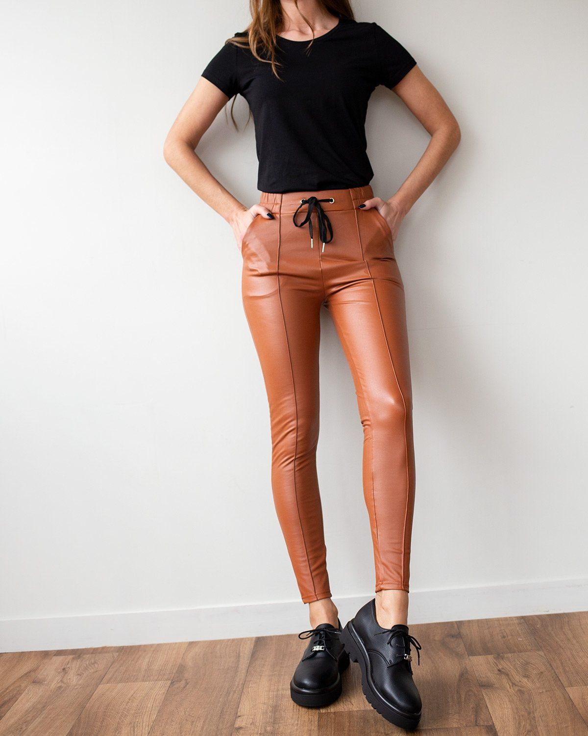 Rust-orange faux leather leggings with black drawstring waist and vertical front seams, slim cropped leg, worn on model, full-length front view