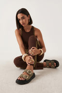 Woman wearing brown outfit and sandals sitting on a white floor.
