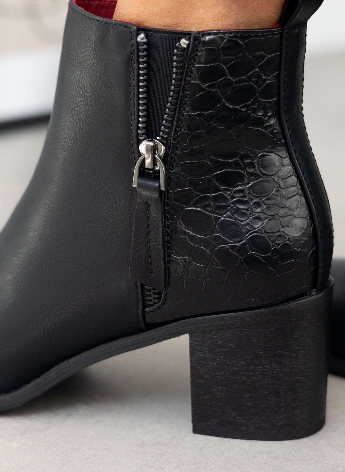 Close-up of croc embossed panel and zipper pull on a black ankle boot with block heel worn on model