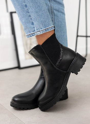Black ankle boots with ribbed elastic panel, glitter trim and chunky lugged sole, worn on model, angled view