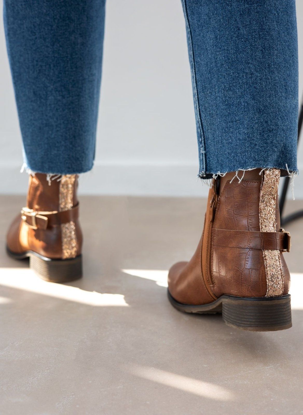 Tan ankle boots with gold buckle strap, side zip and glitter strip at the back of the heel, worn on model, back view