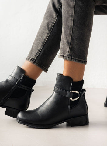 Black faux leather ankle boot with strap and silver ring buckle, low block heel and treaded sole, worn on model, seated view