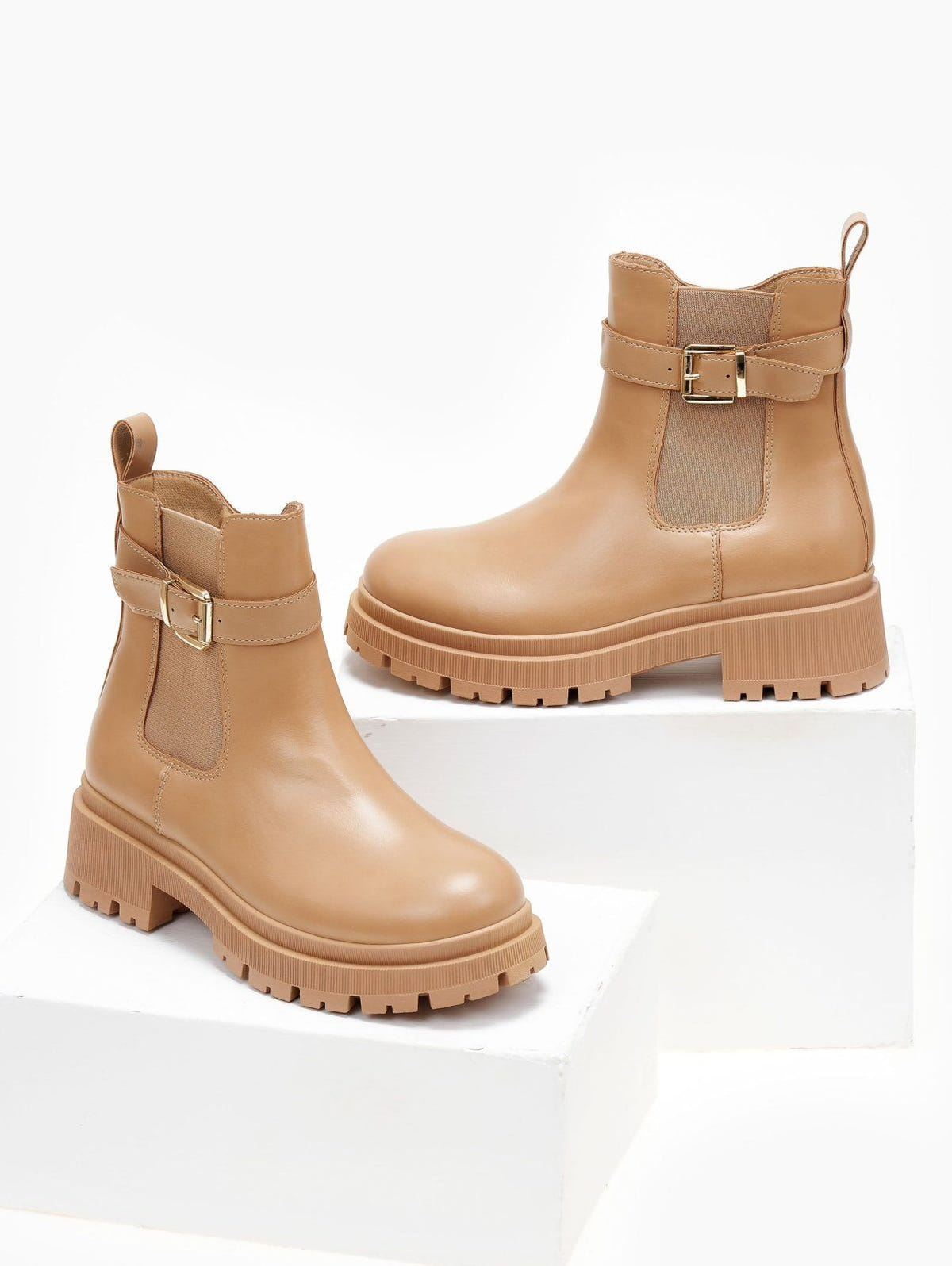 Pair of taupe biker ankle boots with buckle strap, elastic panel and lug platform sole, shown on white blocks
