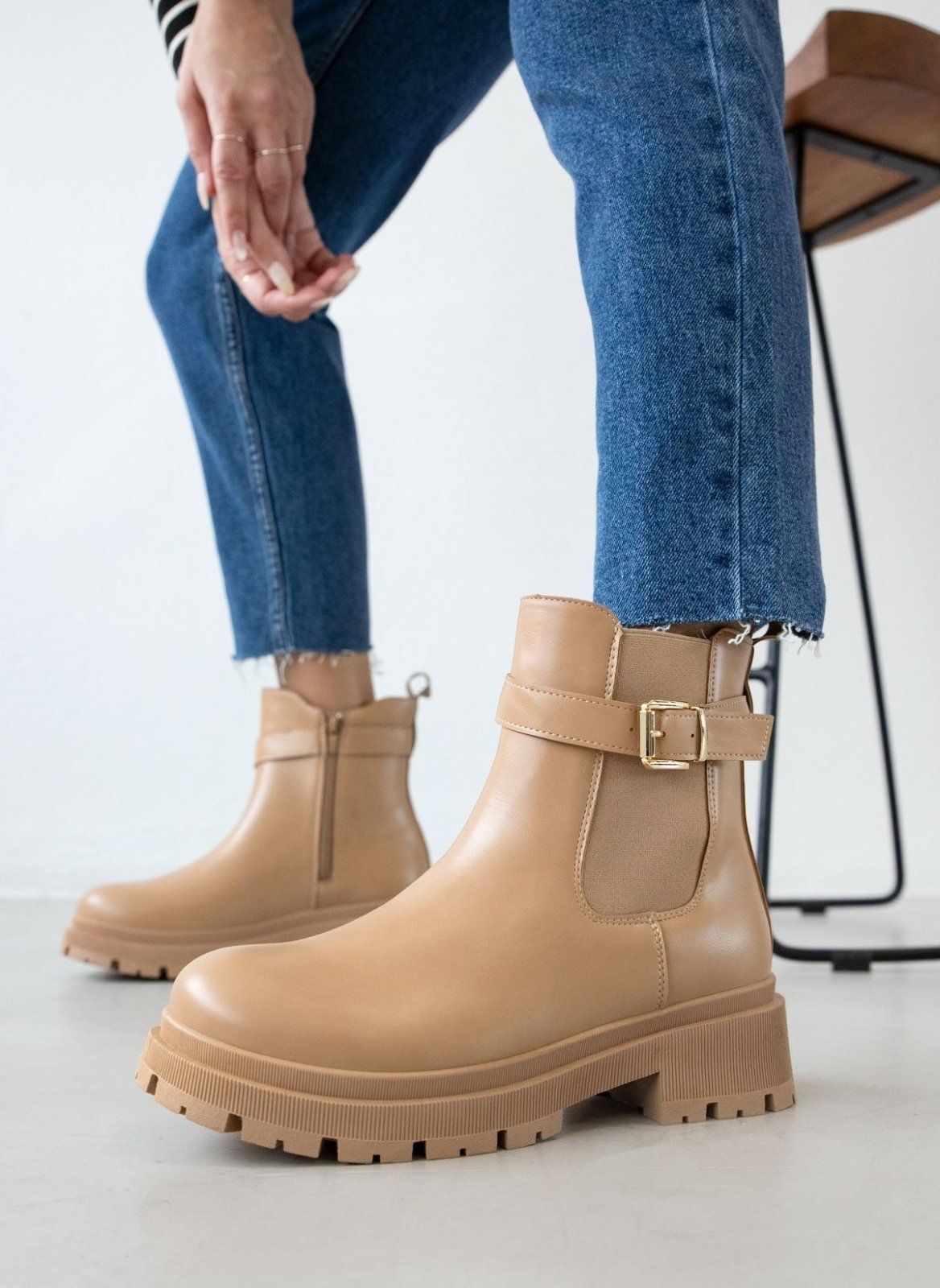 Taupe leather-look biker ankle boots with gold buckle strap, elastic gusset and chunky lug sole, worn on model, side view