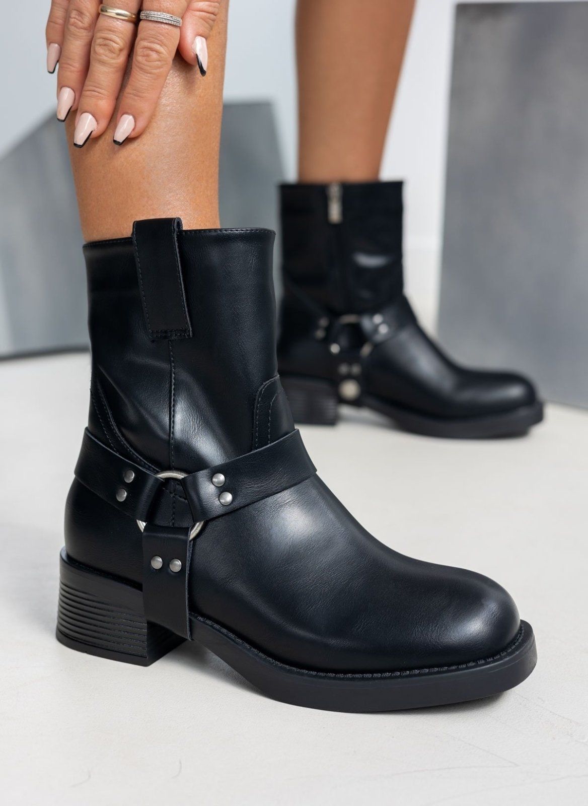 Black faux leather biker boot with metal ring harness strap and block heel, worn on model, side view