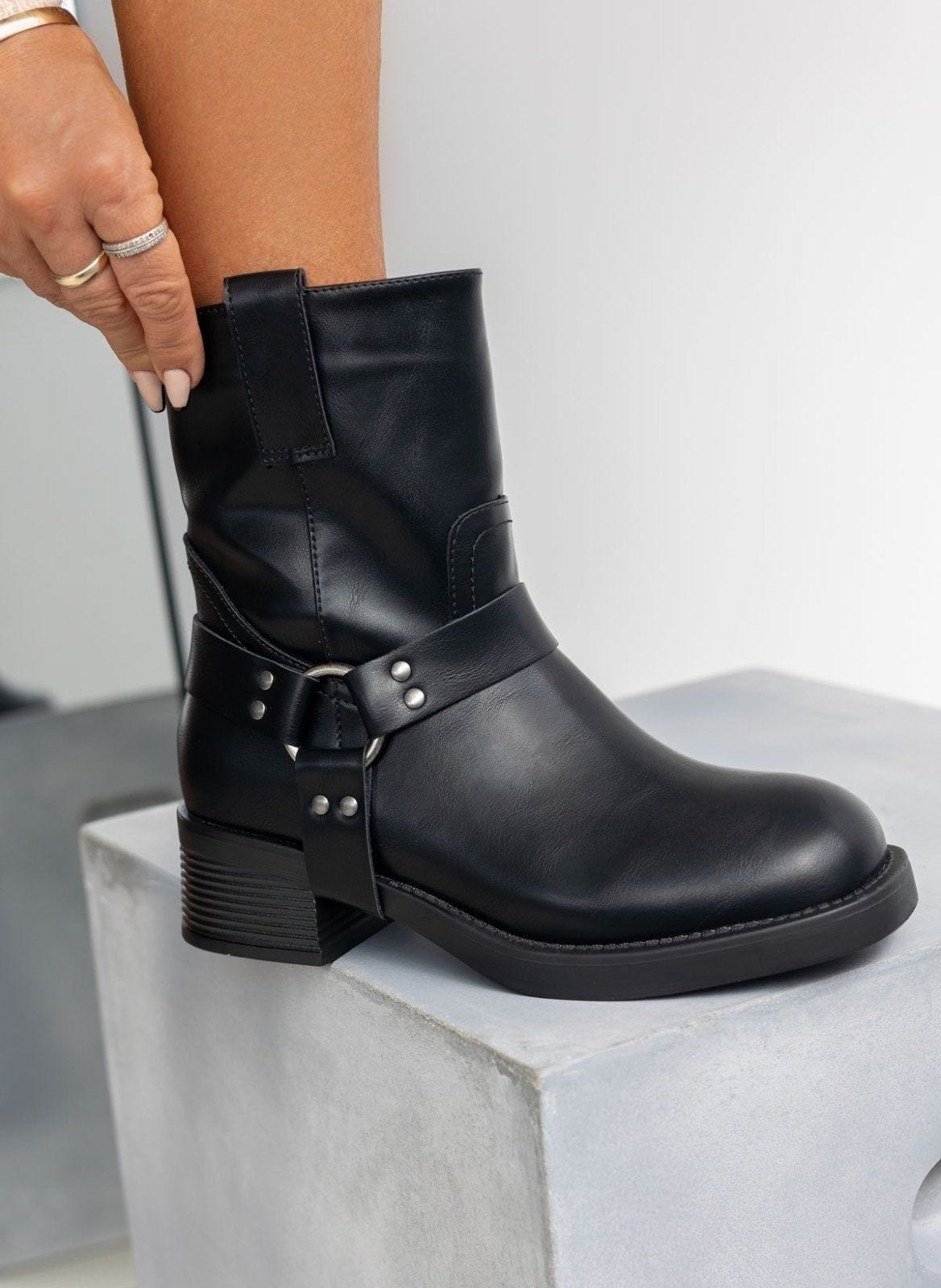 Black faux leather biker boot with silver ring harness strap and block heel, worn on model, resting on a block