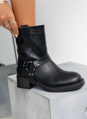Black faux leather biker boot with silver ring harness strap and block heel, worn on model, resting on a block