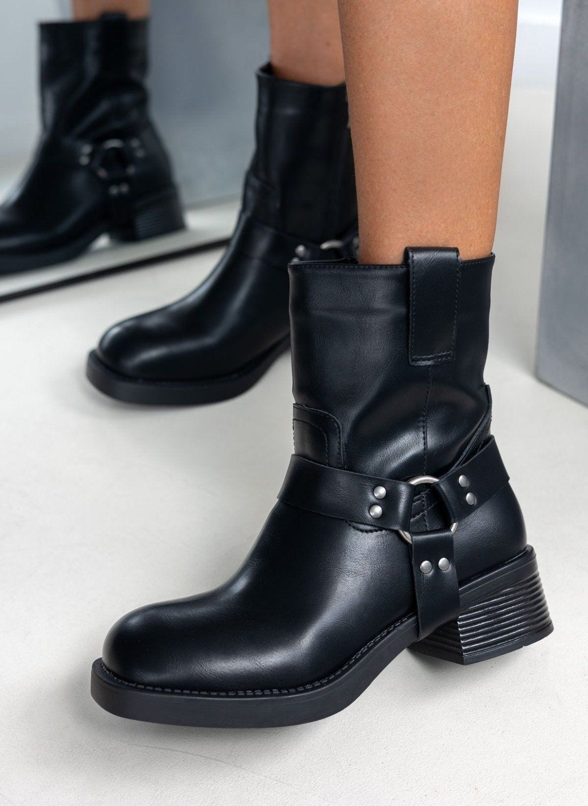 Black faux leather biker boots with ring harness strap and block heel, worn on model, three-quarter view