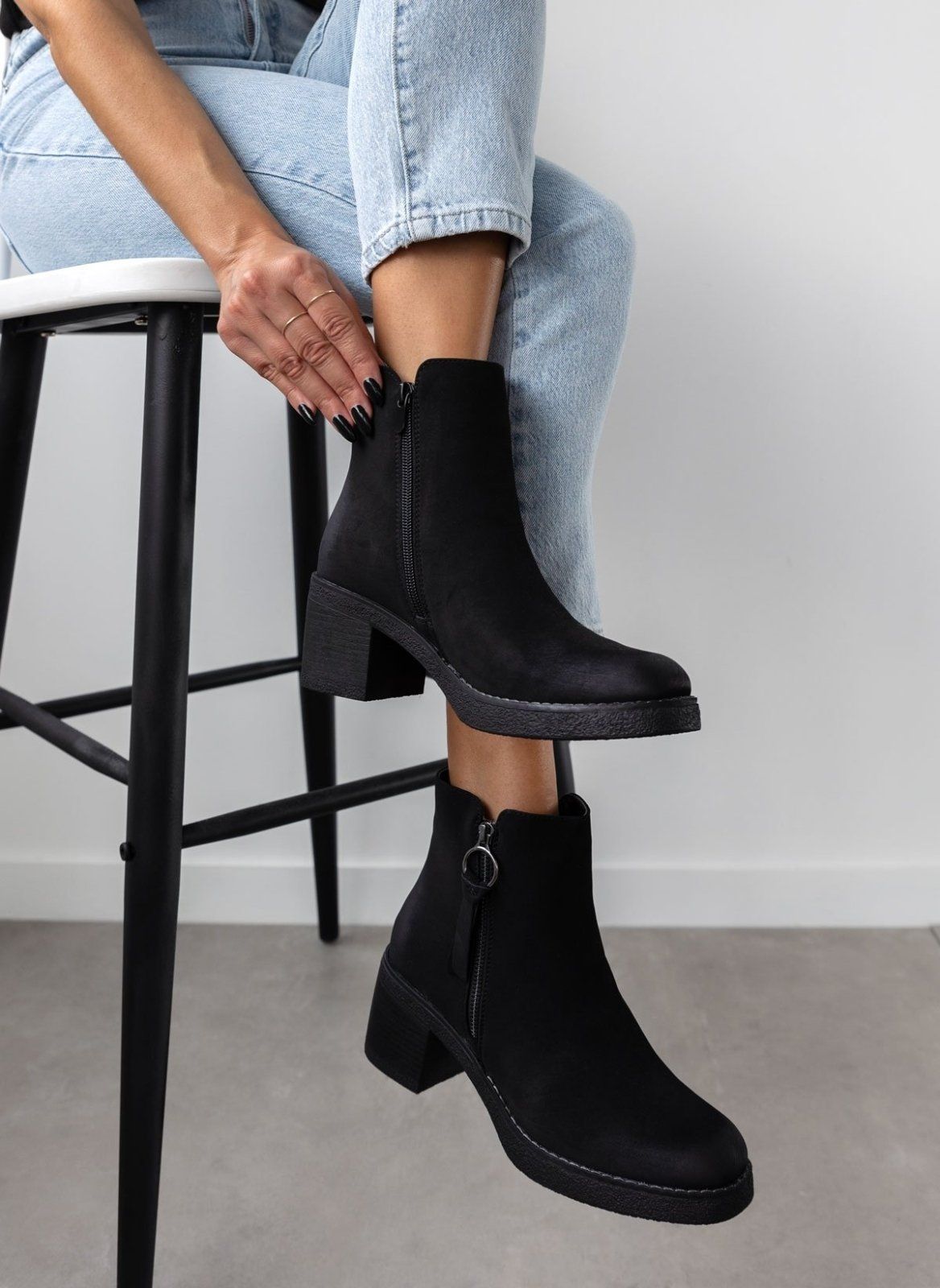 Model seated on a stool wearing black nubuck ankle boots with side zip and block heel, full length