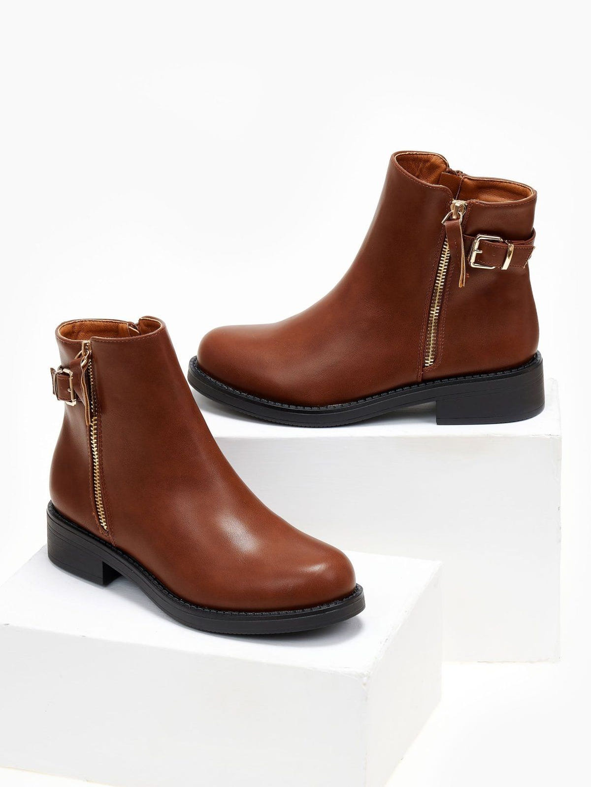 Two tan ankle boots with gold side zippers and buckle straps, displayed on white blocks