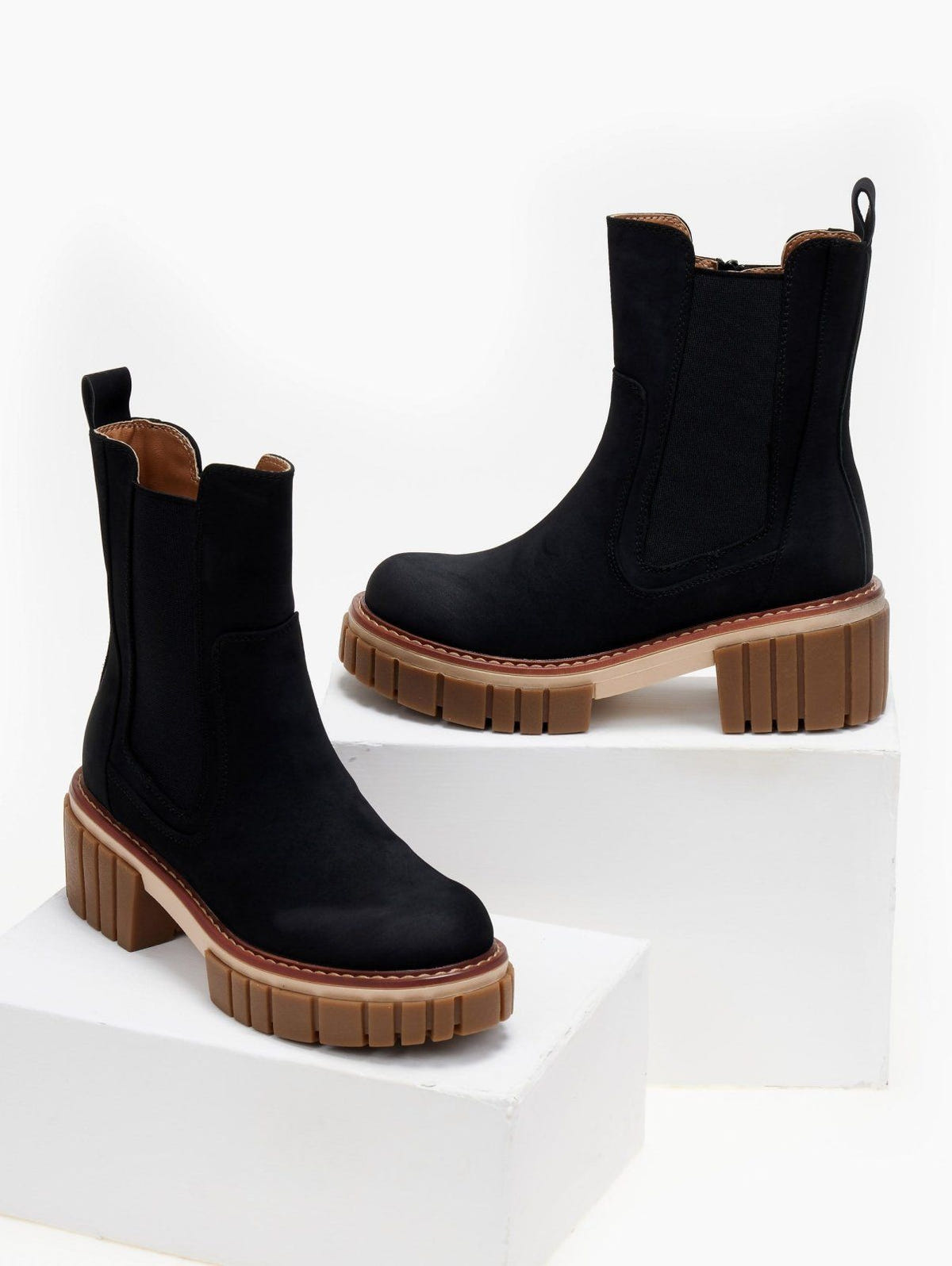 Pair of black suede Chelsea boots with chunky caramel sole, displayed on white blocks, studio view