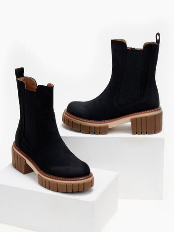 Pair of black suede Chelsea boots with chunky caramel sole, displayed on white blocks, studio view