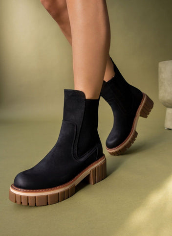 Black suede Chelsea boots with chunky ridged sole and block heel, worn on model against olive backdrop
