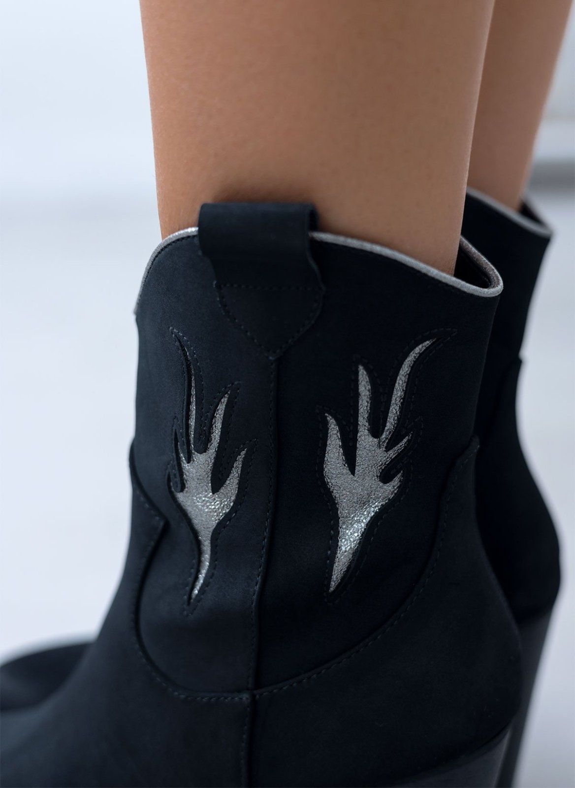 Close-up of silver flame details and silver piping on a black suede cowboy boot worn on model