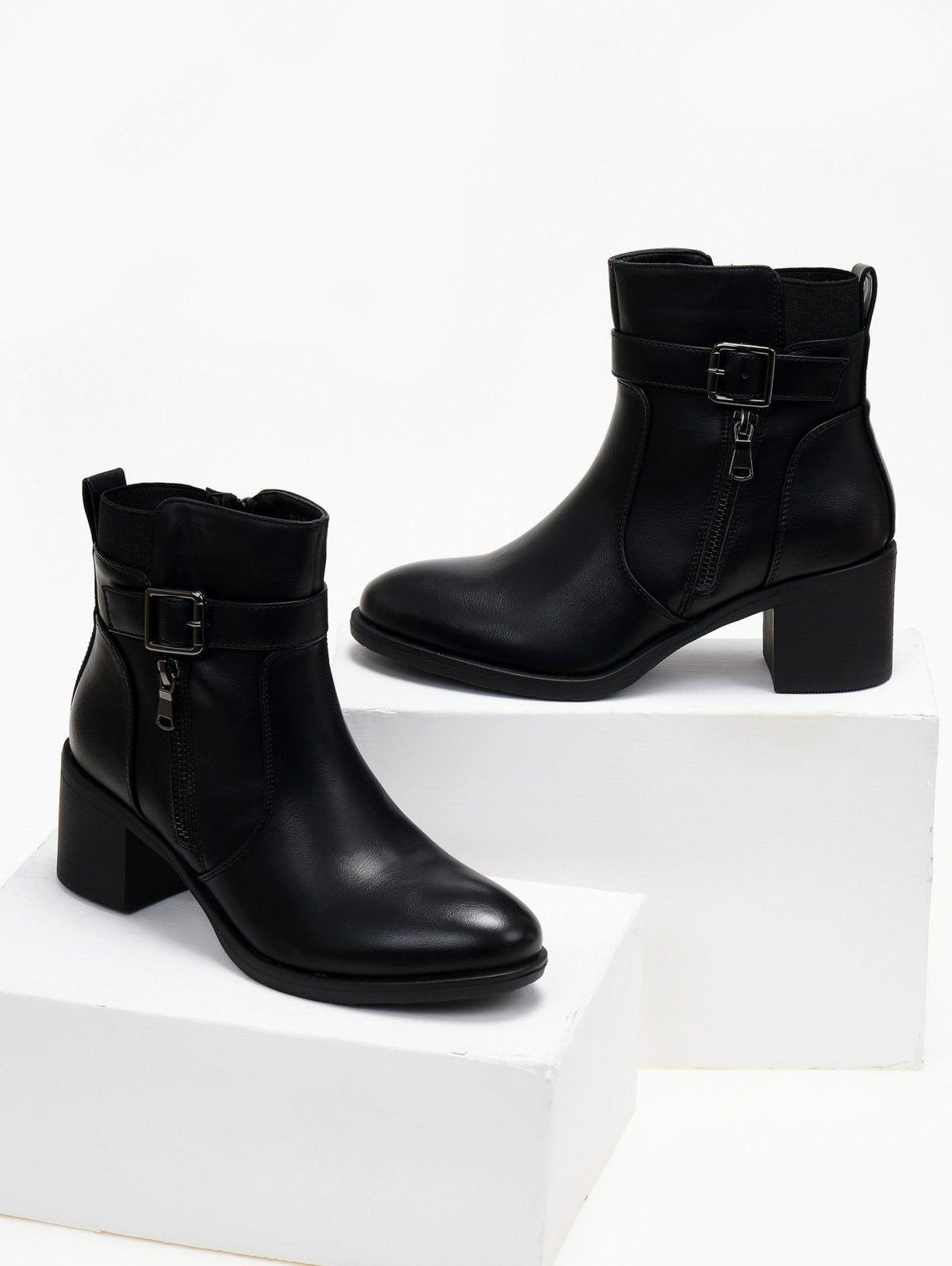 Block heel ankle boots with buckle strap and outer zipper in black, pair displayed on white blocks