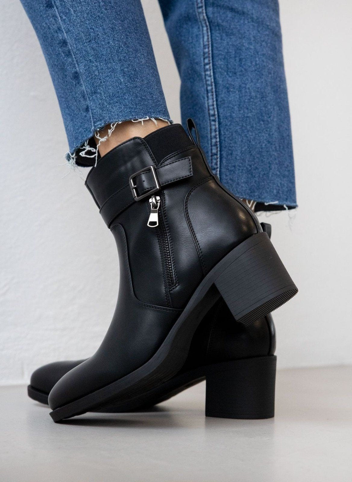 Block heel ankle boots with buckle strap and decorative zipper in black, back side view worn on model