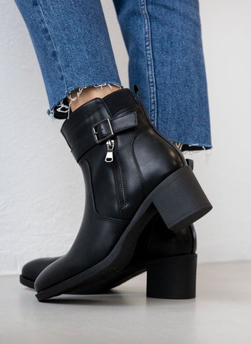 Block heel ankle boots with buckle strap and decorative zipper in black, back side view worn on model