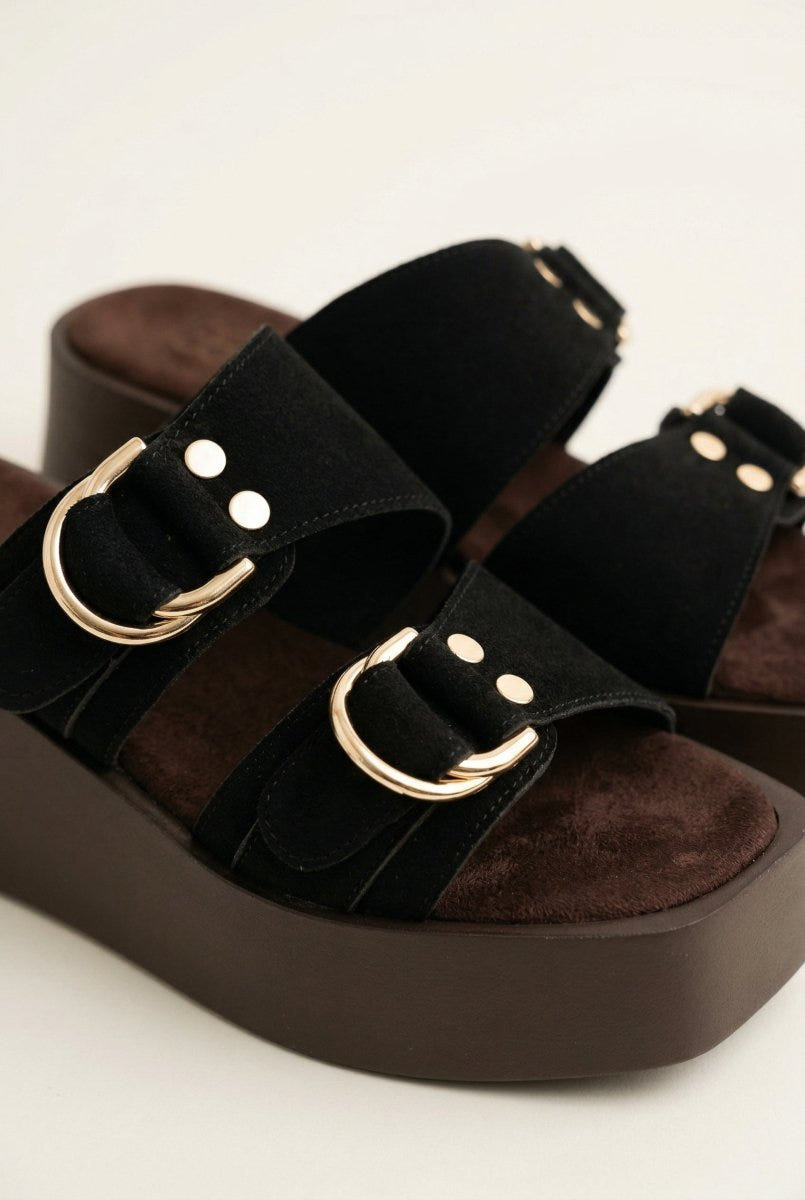 Close-up of black suede strap with gold D-ring buckle and studs over a brown platform sole