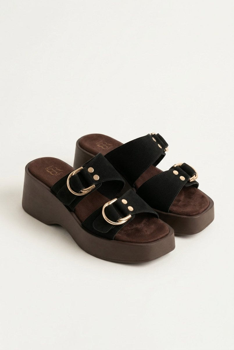 Three-quarter view of black suede platform sandals with gold ring buckles and brown wedge sole, pair on light grey