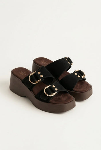 Three-quarter view of black suede platform sandals with gold ring buckles and brown wedge sole, pair on light grey