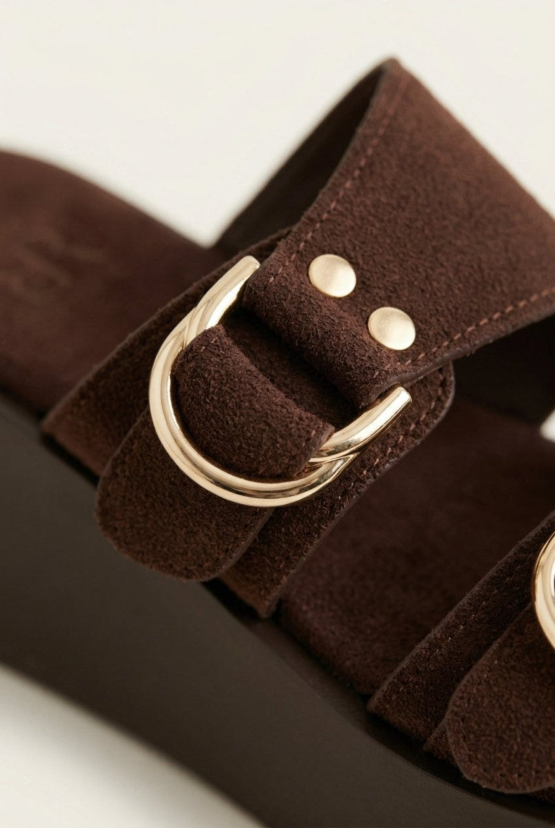 Extreme close-up of brown suede strap with gold ring buckle and small rivets over a platform sole