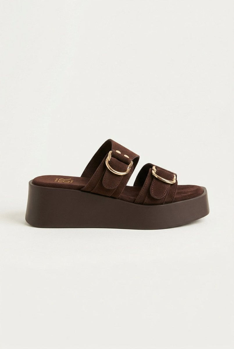 Brown suede platform slide with two straps and gold buckles on thick dark brown sole, side view on light grey