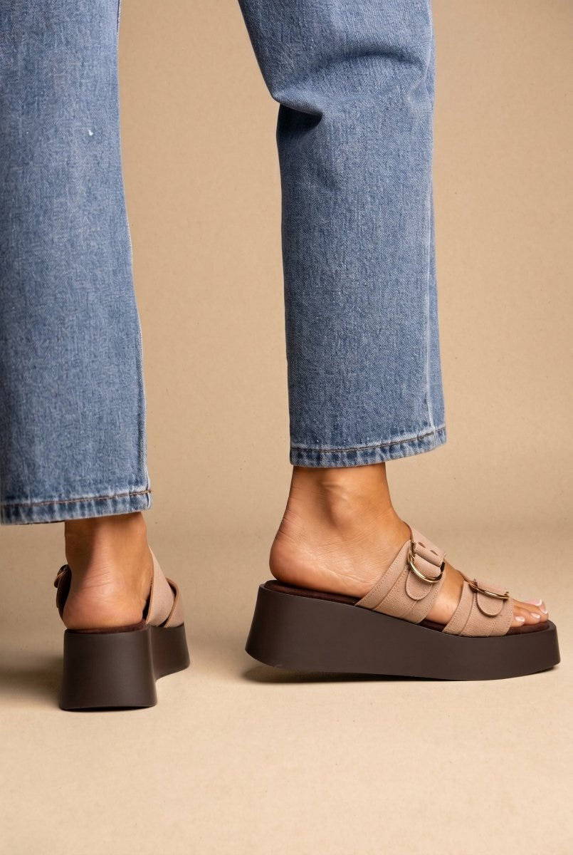Model wearing taupe suede platform sandals with gold buckle straps and blue jeans, side view of feet on beige ground