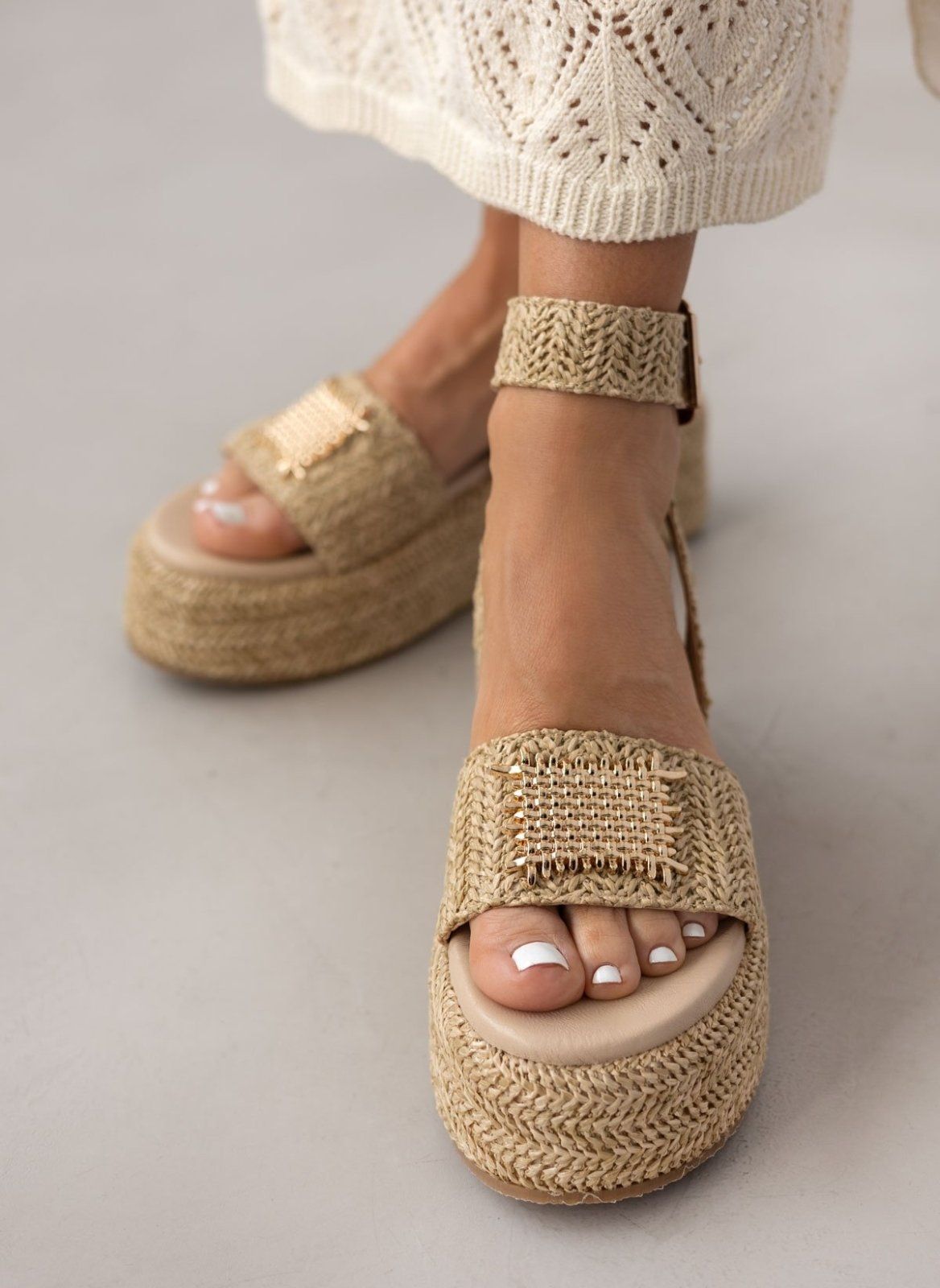 Close-up of model wearing natural raffia platform sandals with gold plate on the wide strap