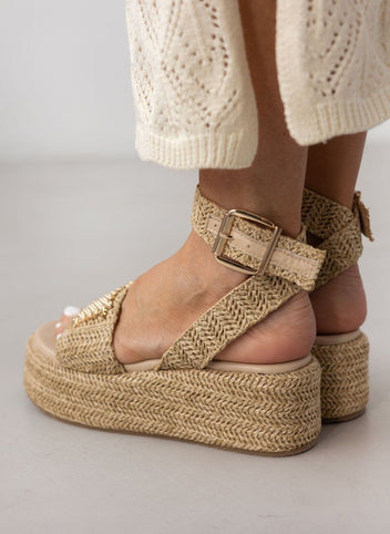 Model wearing natural raffia platform sandals with ankle strap buckle and white crochet trousers, side view